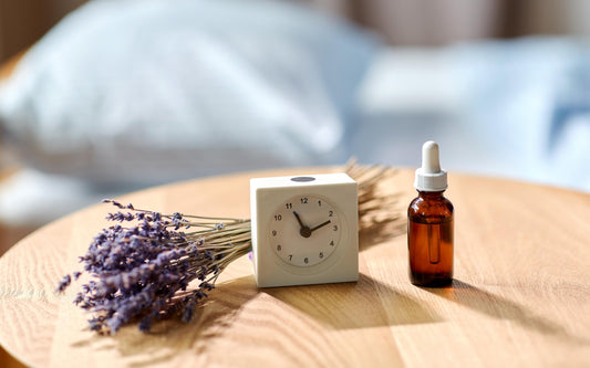 A small white cube-shaped clock showing the time as 10:10, a bunch of dried lavender flowers, and a brown glass dropper bottle are placed on a wooden surface with a blurred bed in the background