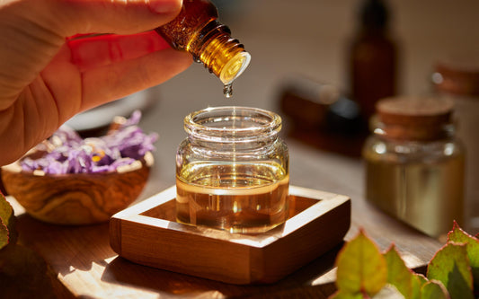 A hand holding a small amber glass bottle, dripping essential oil into a clear glass jar placed on a wooden coaster. In the background, there are dried lavender flowers in a wooden bowl and other small bottles.