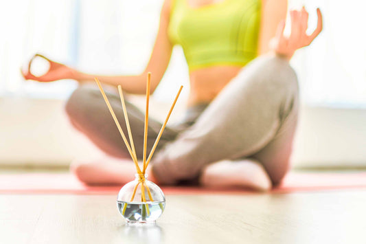 A person sitting cross-legged on a yoga mat in a meditative pose with hands resting on knees, fingers forming a mudra. In the foreground, there is a small glass container with several incense sticks placed inside it.