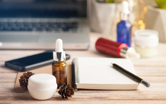 Workspace with skincare items and tech gear on a wooden surface, including a white jar, brown dropper bottle, notebook with pen, pine cones, smartphone, and laptop, set against a cozy background with plants and warm lighting.