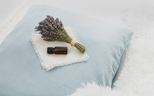 Light blue pillow on a fluffy white surface with dried lavender, a small brown glass bottle, and a fringed white fabric square arranged on top
