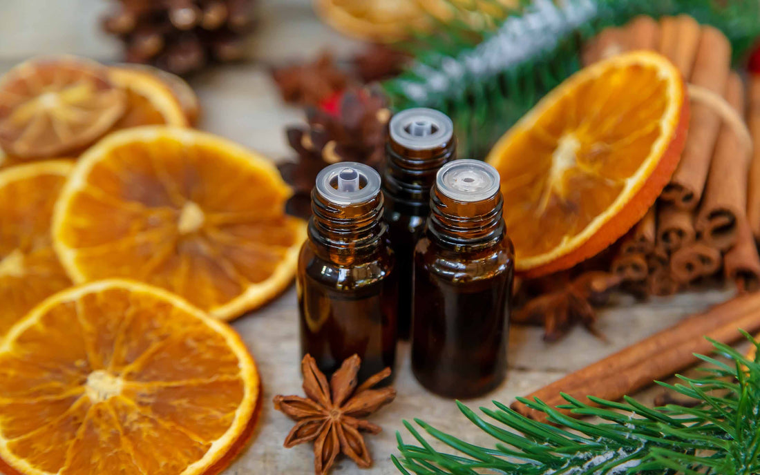 Three small amber glass bottles surrounded by dried orange slices, cinnamon sticks, star anise, pinecones, and evergreen branches in a festive, aromatic arrangement