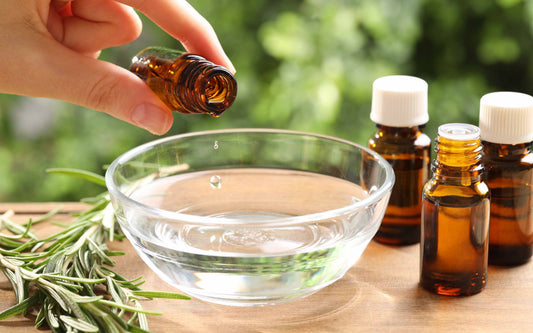 A hand drops oil from a small amber bottle into a clear bowl of water, with other bottles and rosemary sprigs nearby on a wooden surface against a blurred green background.