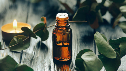 Brown glass bottle of essential oil on a wooden surface with eucalyptus leaves and a lit candle.