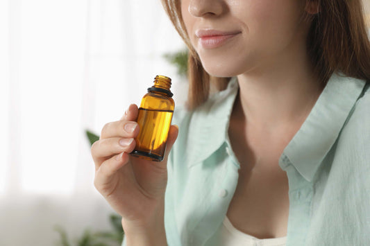 Person holding a small amber glass bottle close to the face, wearing a light blue shirt