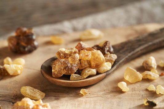 A wooden spoon filled with various sizes of amber-colored resin pieces, placed on a wooden surface. Additional resin pieces are scattered around the spoon on the surface. The background is blurred, highlighting the texture and color of the resins.